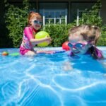 Two kids wearing swim goggles and pink rash guards play with colorful balls at the edge of a blue pool. Lush green bushes and a white railing are visible in the background.