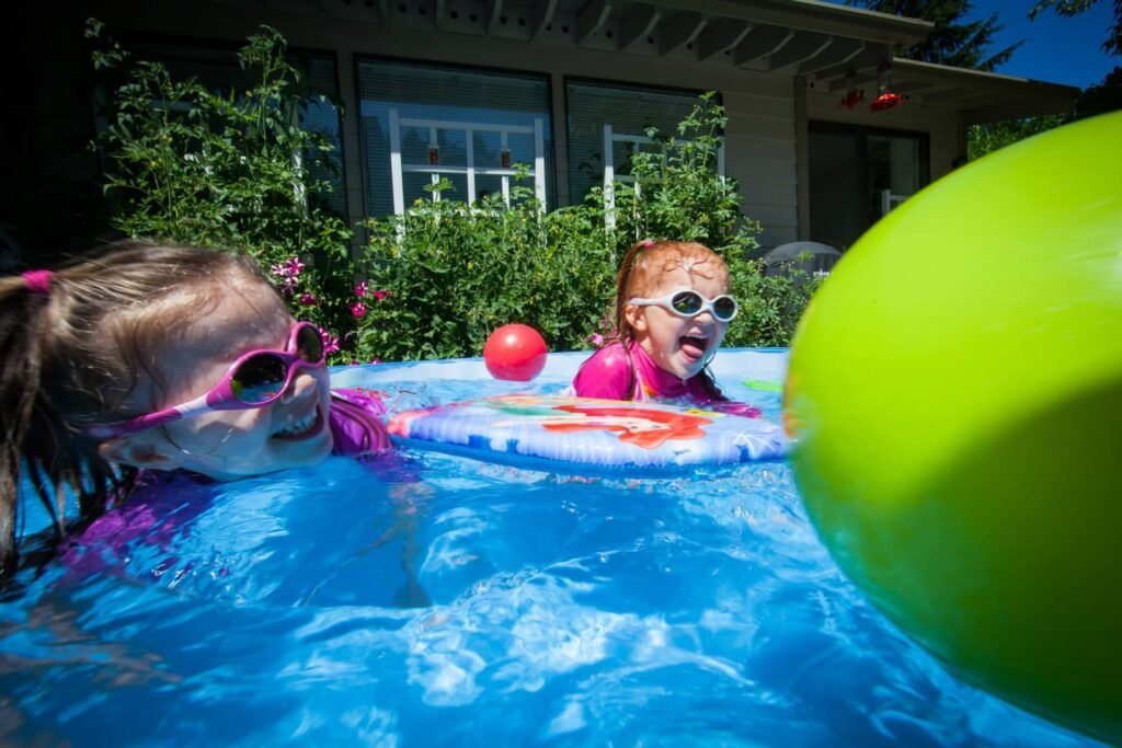 Children playing in a bright blue swimming pool with colorful inflatables and a large green float. A house with windows and garden foliage is visible in the background.