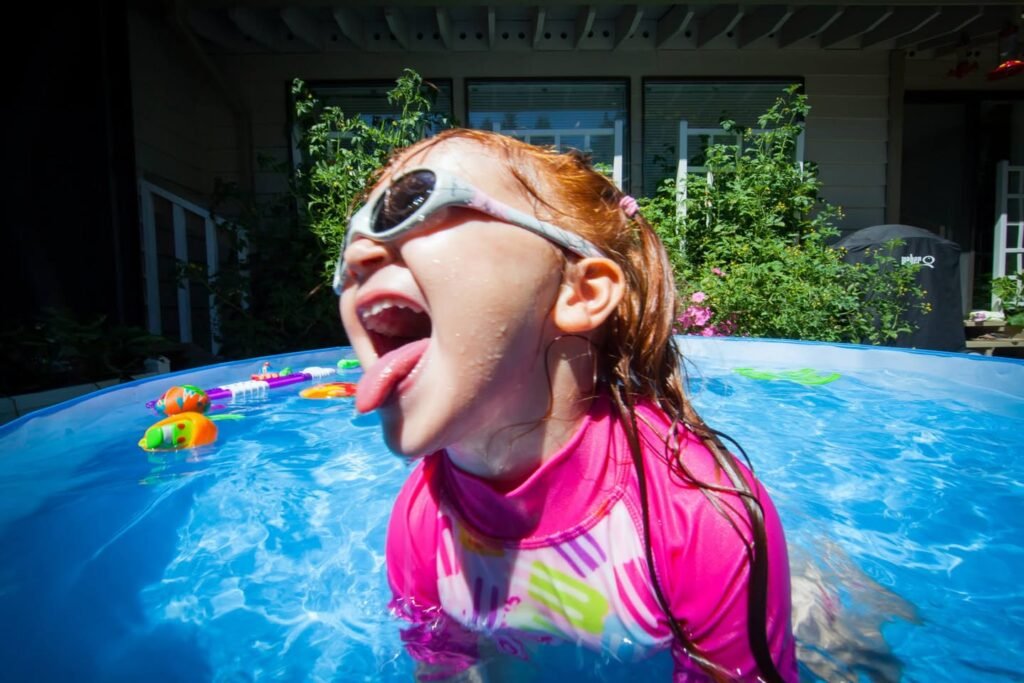 A person in a pink swim shirt and white goggles laughing in a pool, captured with a fisheye lens near a house.