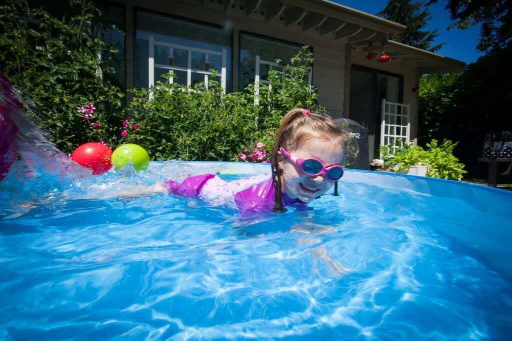 A child plays in a backyard pool with bright balls and greenery surrounding the area.