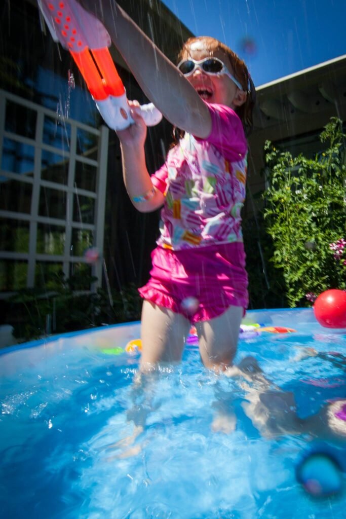 A child making a splash in a pool while holding a red and white water gun.
