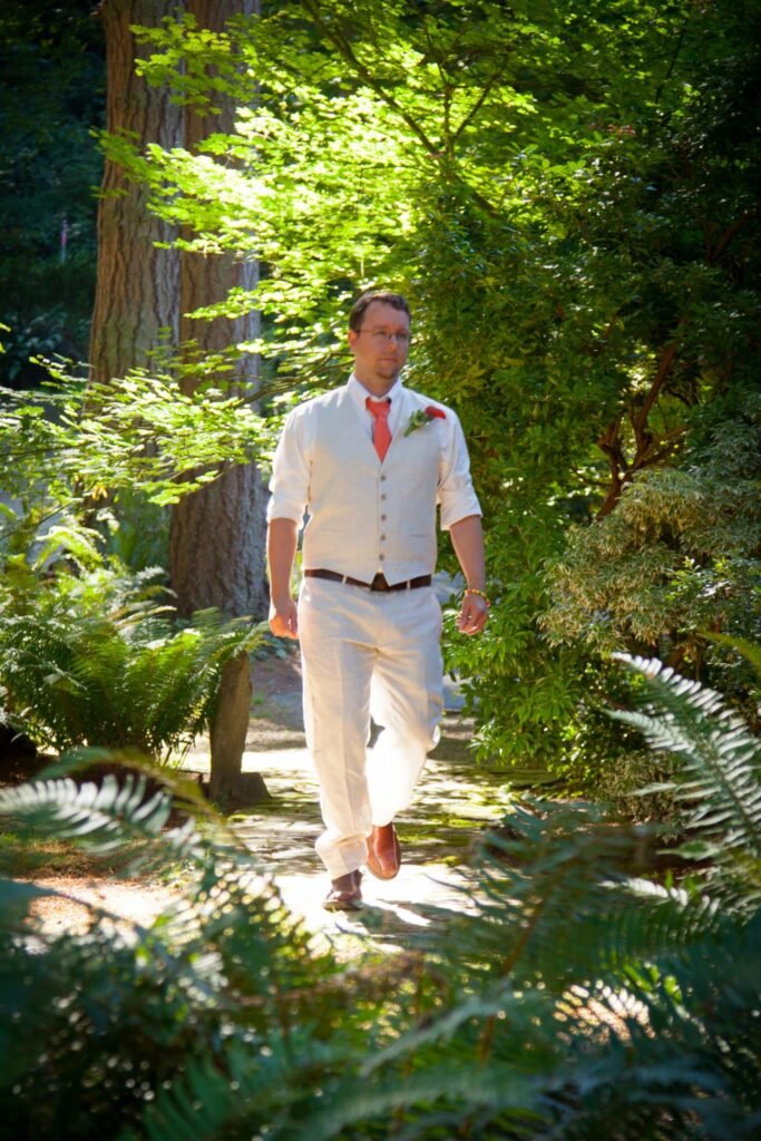 A man in a white wedding outfit walks along a forest path surrounded by ferns and trees.