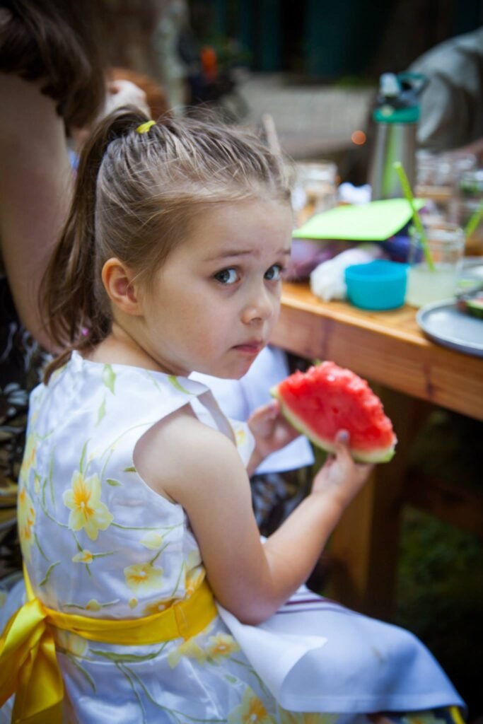 A young girl in a floral dress holding a slice of watermelon at a table outdoors.
