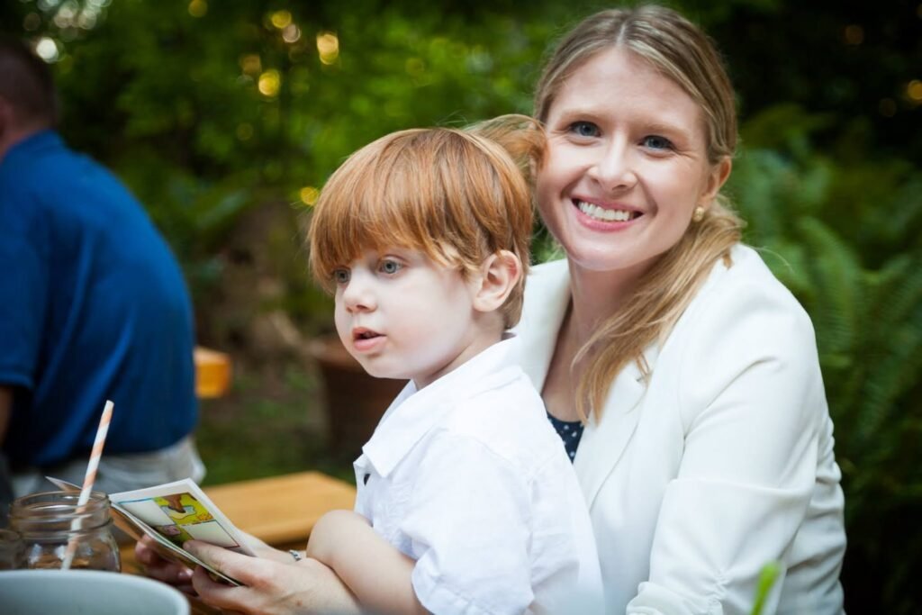 A woman and a child sit outdoors at a table, smiling amid green foliage.