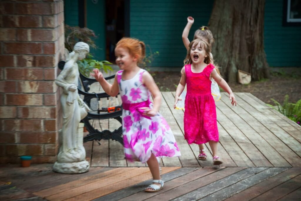 Two young girls wearing pink and white floral dresses run happily across a wooden deck.