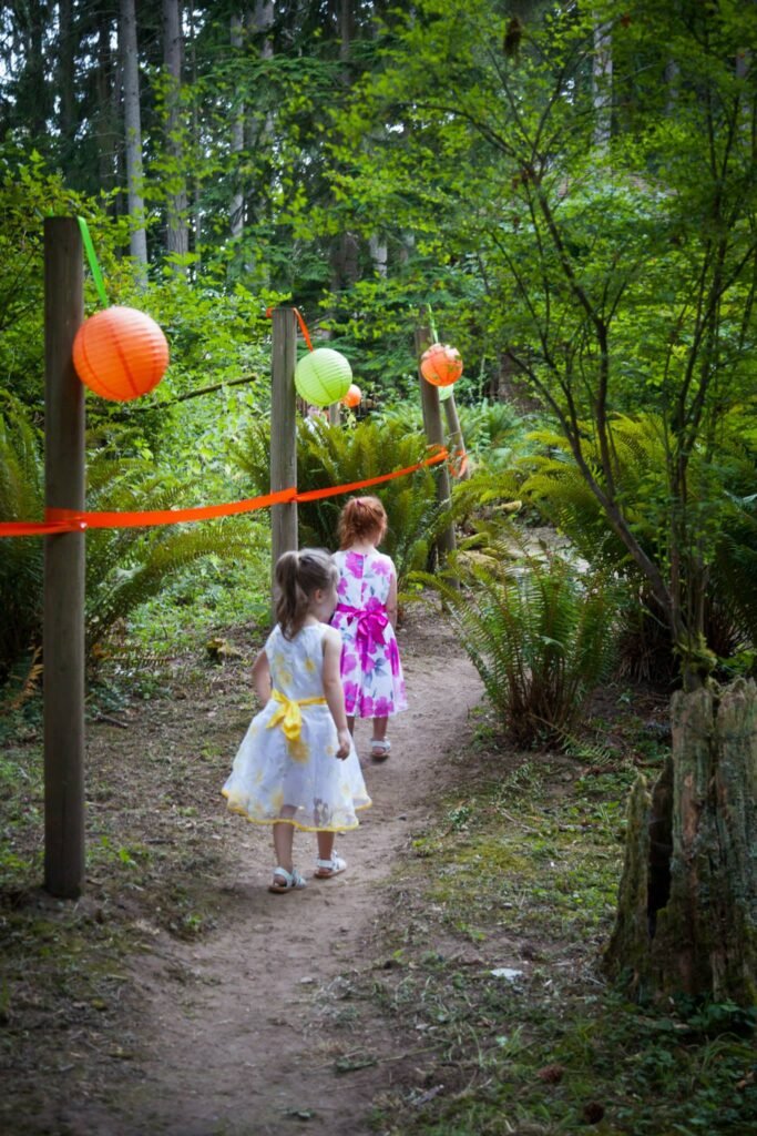 A dirt path in a lush green forest leads past tall evergreens and wooden posts decorated with colorful balloons.