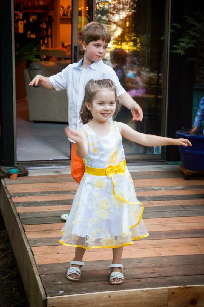A child in a white and yellow dress stands on a wooden deck with arms outstretched, with another child behind them.