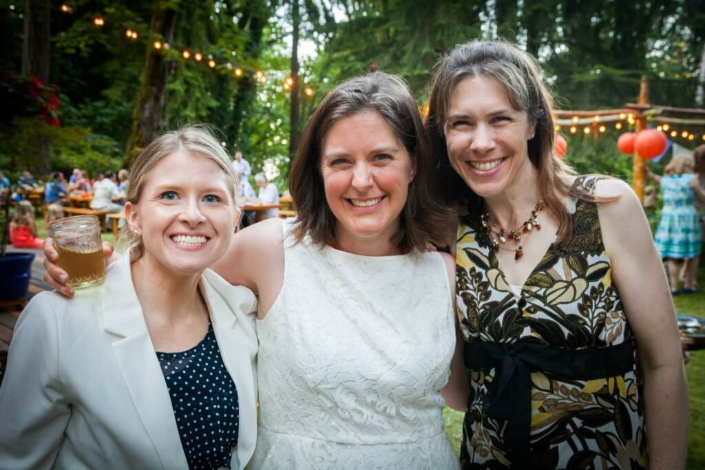 Three friends smiling at an outdoor wedding ceremony with string lights and lush foliage.