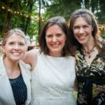 Three friends smiling at an outdoor wedding ceremony with string lights and lush foliage.