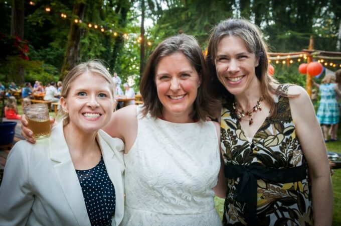 Three friends smiling at an outdoor wedding ceremony with string lights and lush foliage.