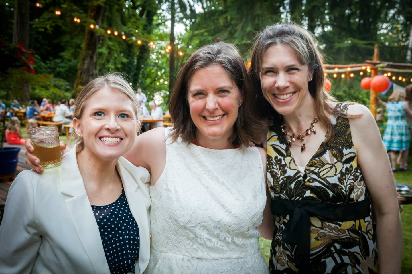 Three friends smiling at an outdoor wedding ceremony with string lights and lush foliage.