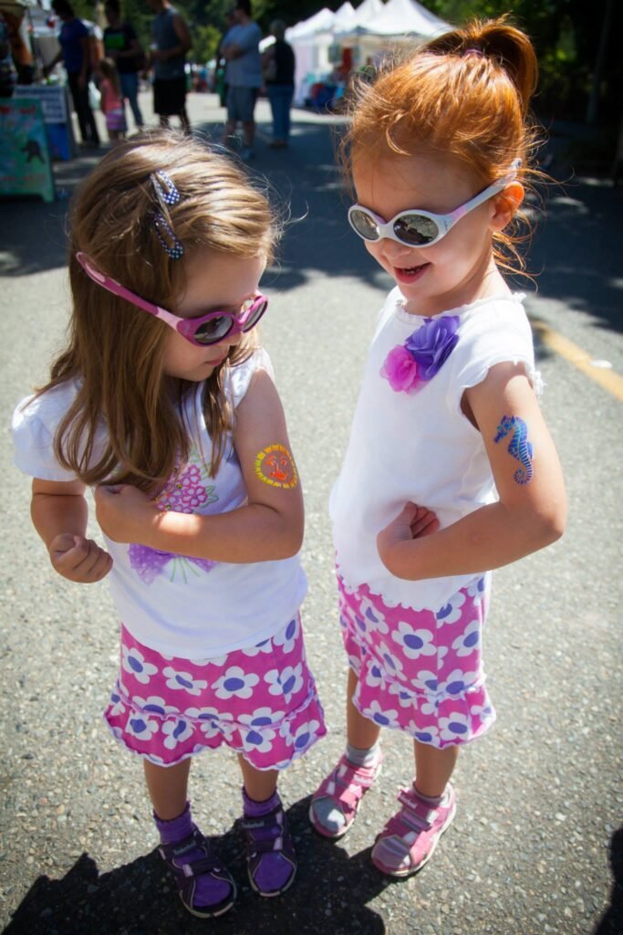 Two young children wearing sunglasses and matching pink tops and dresses stand outside with face paint visible.