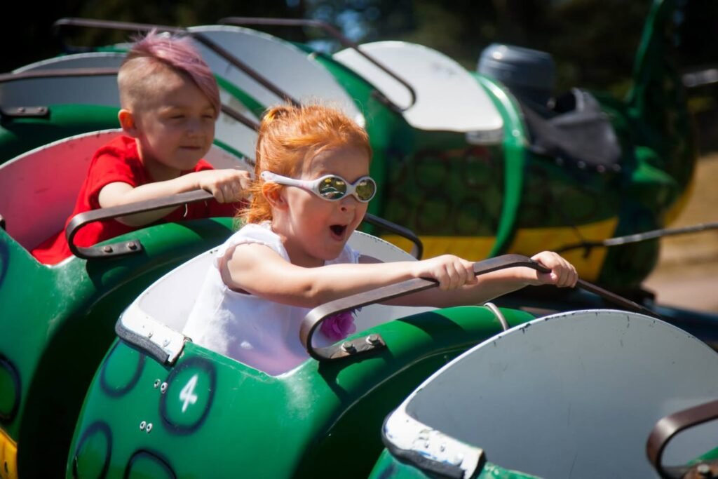 Two children ride a green roller coaster with one wearing sunglasses and the other in a red shirt.