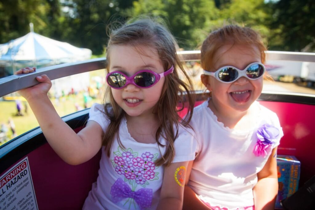Two children wearing colorful sunglasses sit in a red vehicle at an outdoor event. A white circus tent is visible in the background.