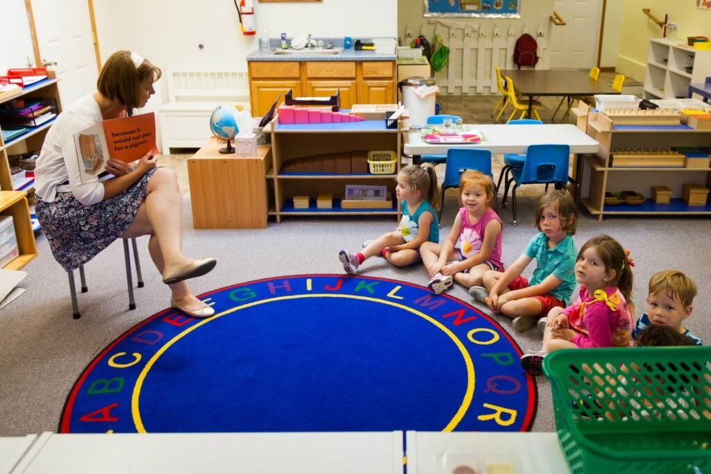 A classroom setup featuring a blue circular rug with alphabet letters, a seated adult, and a group of young children. Educational furniture and materials are visible in the background.