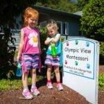 Two children in colorful clothes stand beside a sign for the Olympic View Montessori preschool in a sunny, landscaped area.