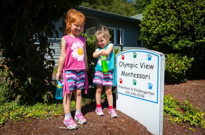 Two children in colorful clothes stand beside a sign for the Olympic View Montessori preschool in a sunny, landscaped area.