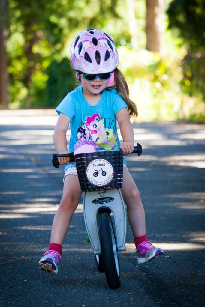 A child in a blue shirt and pink helmet riding a white balance bike outdoors.