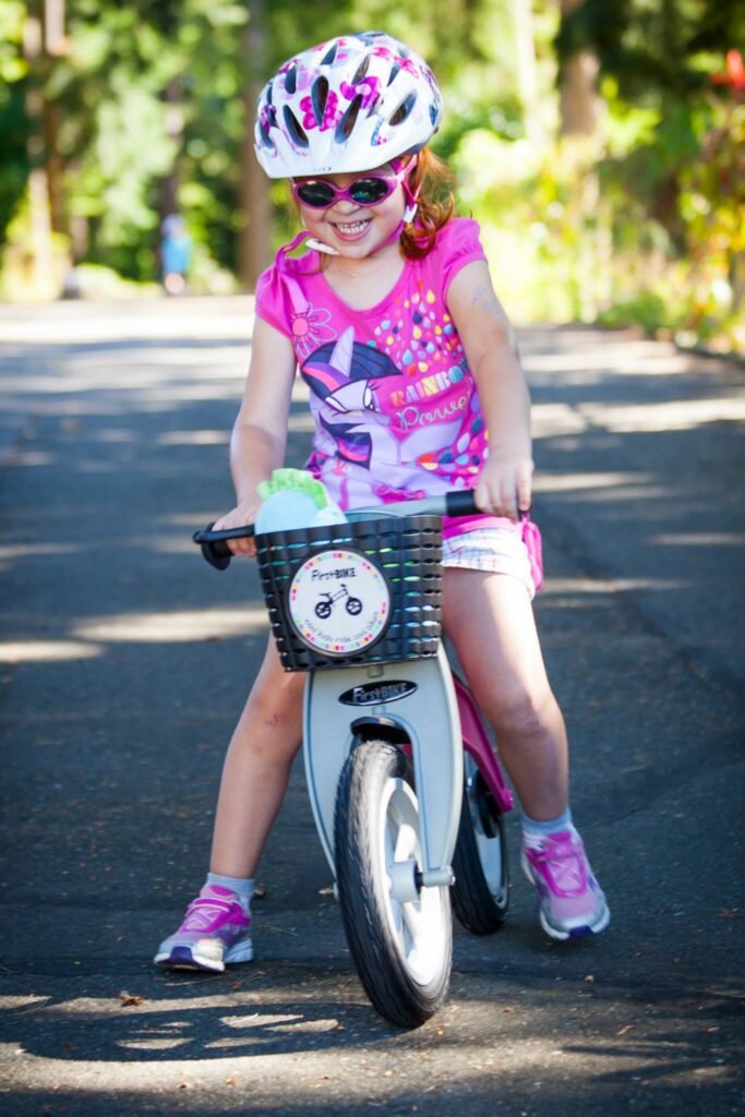 A child in a white helmet and pink bicycle outfit rides a white balance bike on a paved outdoor path. The bike features a front basket and sporty pink shoes complete the look.