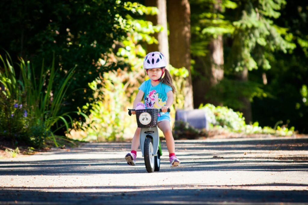 A young child riding a balance bike on a paved path through a lush, wooded area. The child is wearing a white helmet and a blue shirt.
