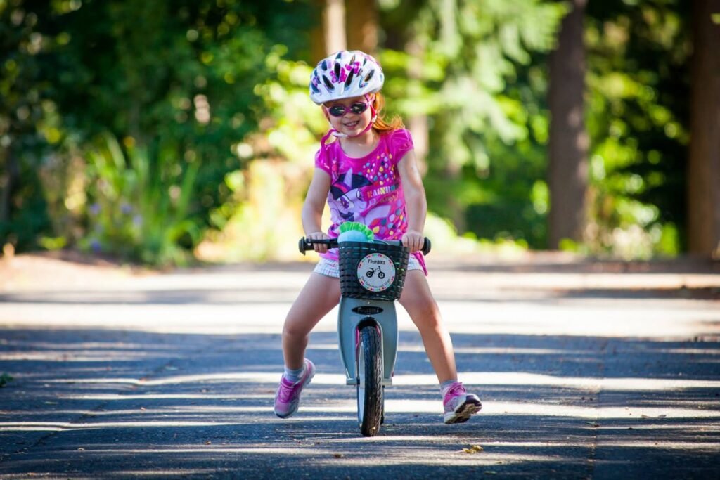 A child wearing a helmet and sunglasses rides a small bike on a paved path. The background features lush greenery and dappled sunlight.