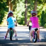 Two children in colorful clothing and protective helmets ride bicycles along a sunny paved path.