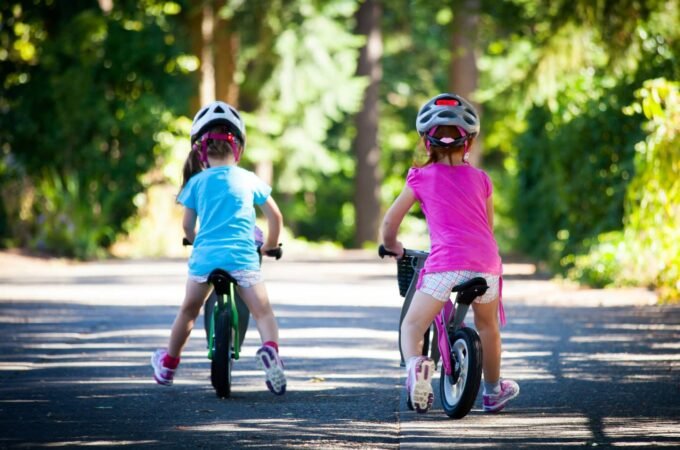 Two children in colorful clothing and protective helmets ride bicycles along a sunny paved path.