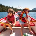 Two children row a red boat on a calm lake with a forested shoreline and a distant mountain in the background. A second view shows the surrounding forest and mountainscape, highlighting the natural beauty of the trip.
