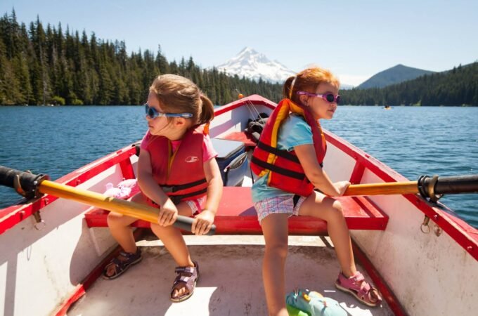 Two children row a red boat on a calm lake with a forested shoreline and a distant mountain in the background. A second view shows the surrounding forest and mountainscape, highlighting the natural beauty of the trip.
