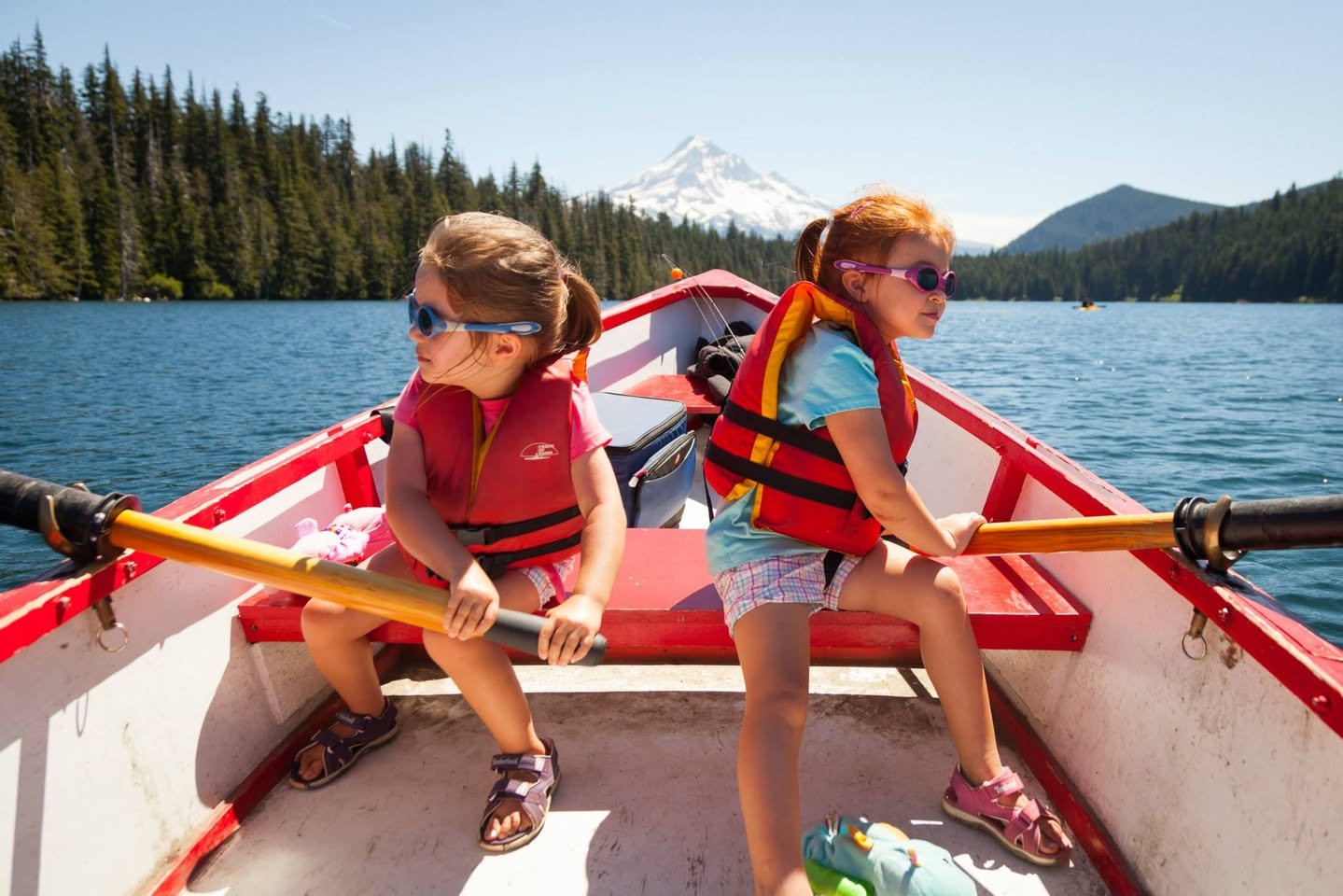 Two children row a red boat on a calm lake with a forested shoreline and a distant mountain in the background. A second view shows the surrounding forest and mountainscape, highlighting the natural beauty of the trip.