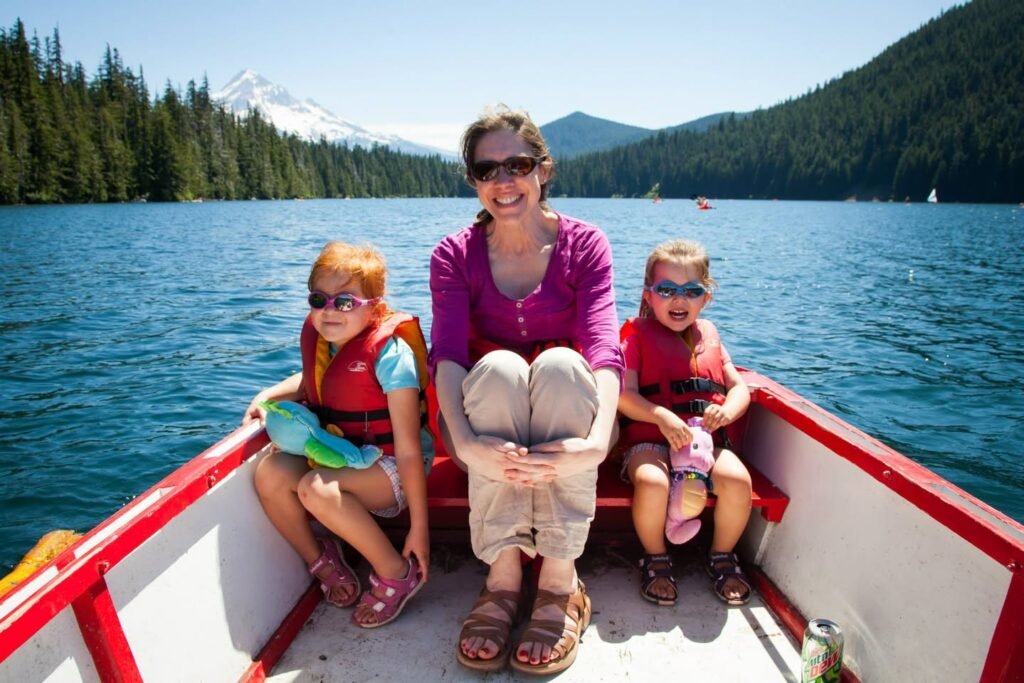 A smiling woman and two children wearing life jackets sitting in a red and white boat at a scenic lake with forested mountains in the background.