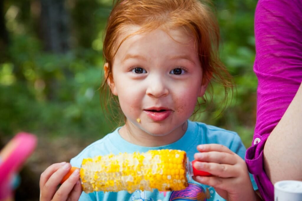 A child holds a cob of buttered corn outdoors with a blurred green background.