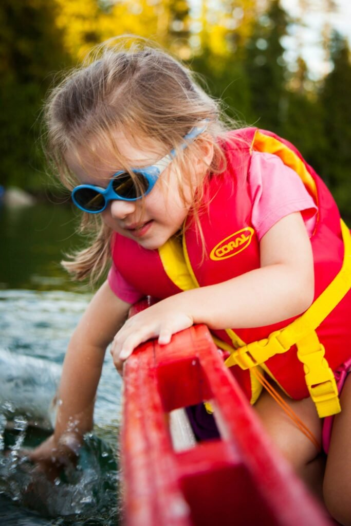 A child wearing a red life jacket and blue sunglasses plays at the edge of a body of water. A second close-up shows the child holding a red float and touching the water.