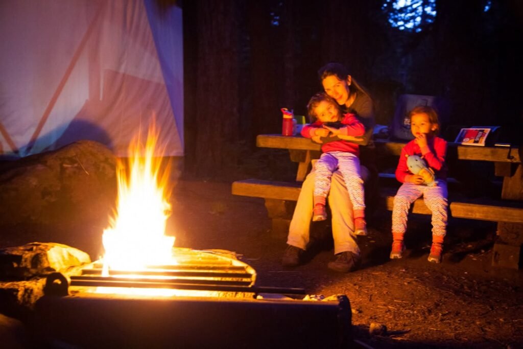 A family gathers around a campfire at night near a tent and picnic table, enjoying a cozy camping trip. The fire provides a warm light in the dark forest setting.