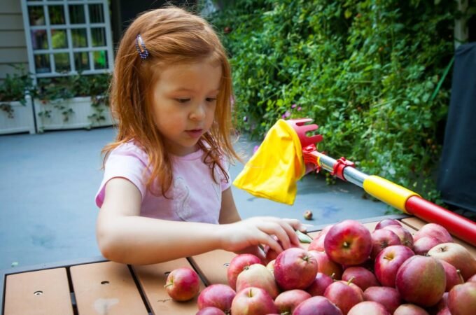 A young child carefully selects apples from a wooden table outdoors, with a colorful toy nearby.