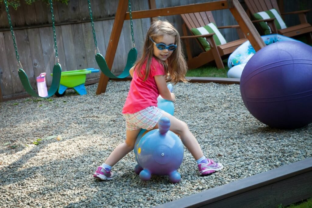 A girl riding a purple inflatable turtle ball in a gravel play yard. Green swings and a purple exercise ball are visible in the background.