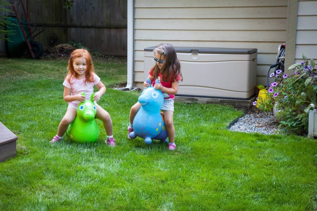 Two children play with large green and blue garden sprinkler toys on a backyard lawn. A patio table with hanging planter and flowers is visible nearby.