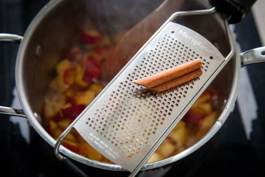 Cinnamon sticks rest on a metal strainer over a pot of simmering apple cider.