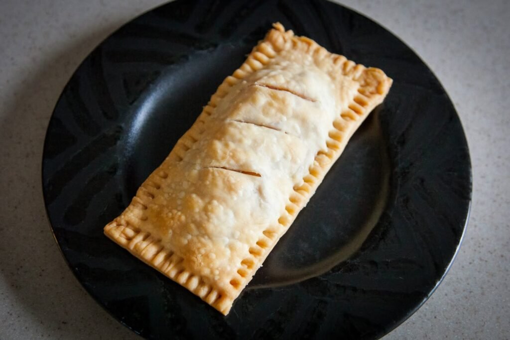 A golden-brown beef wellington with decorative crimped edges is served on a dark plate.