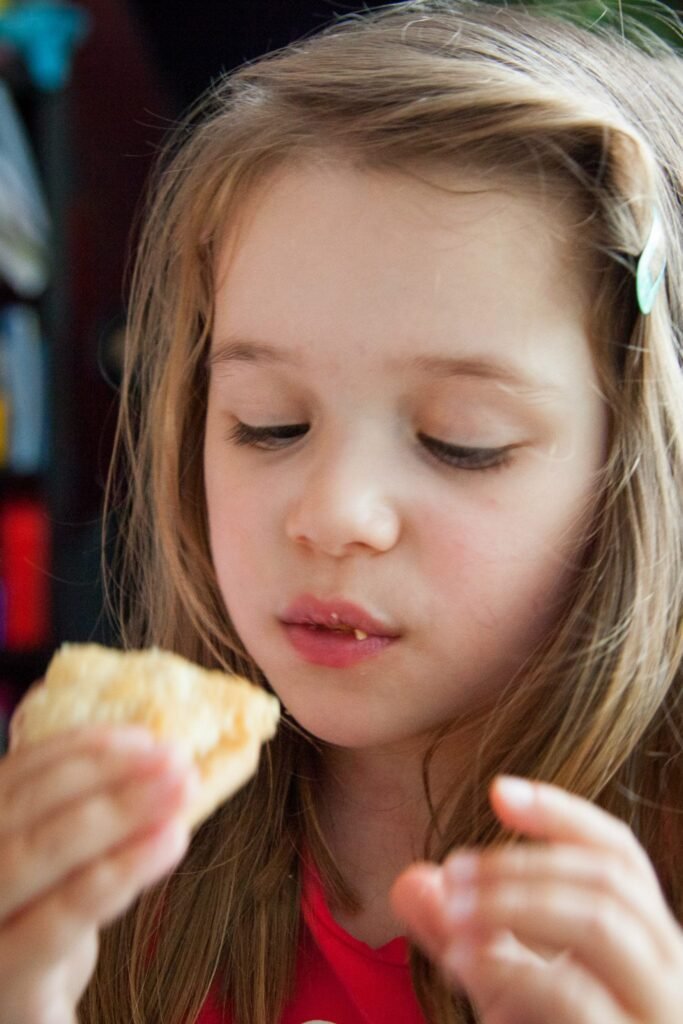 A young child with long blonde hair in a red shirt is shown eating a light food item.