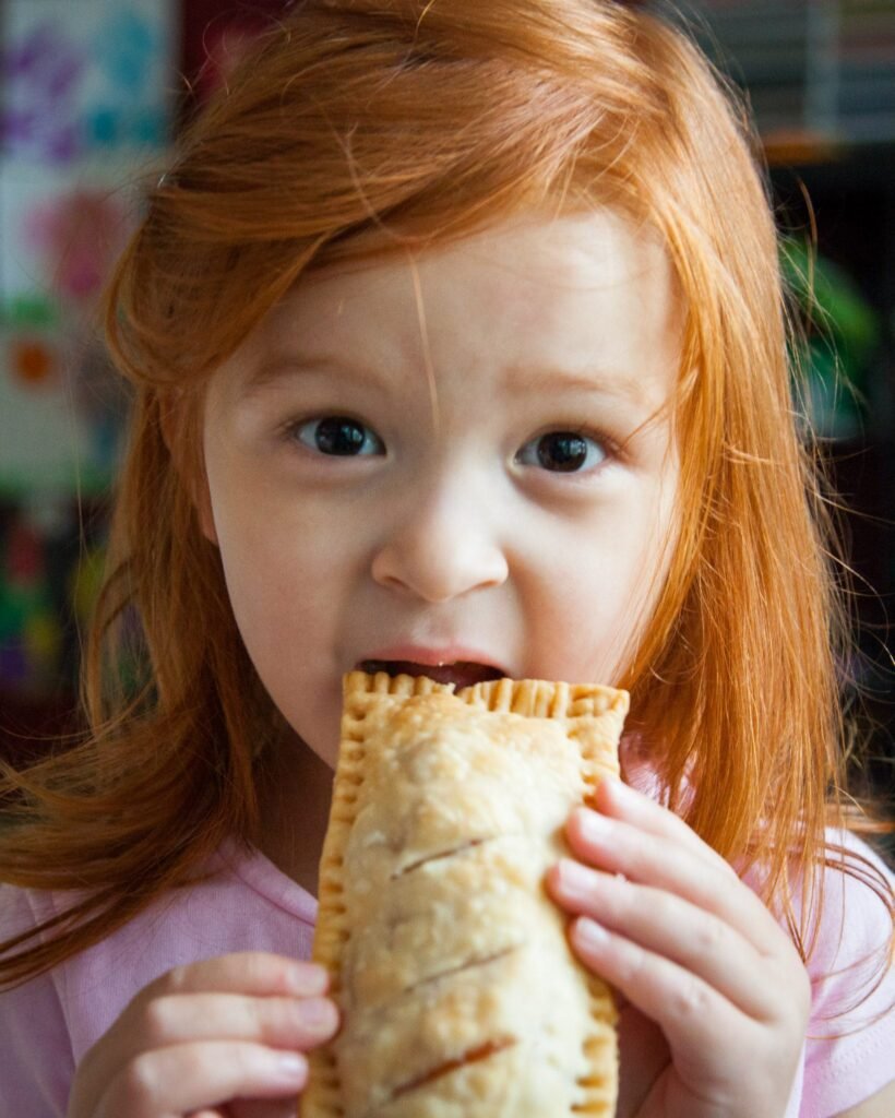 A young child with red hair takes a bite from a golden-brown crimped pie.