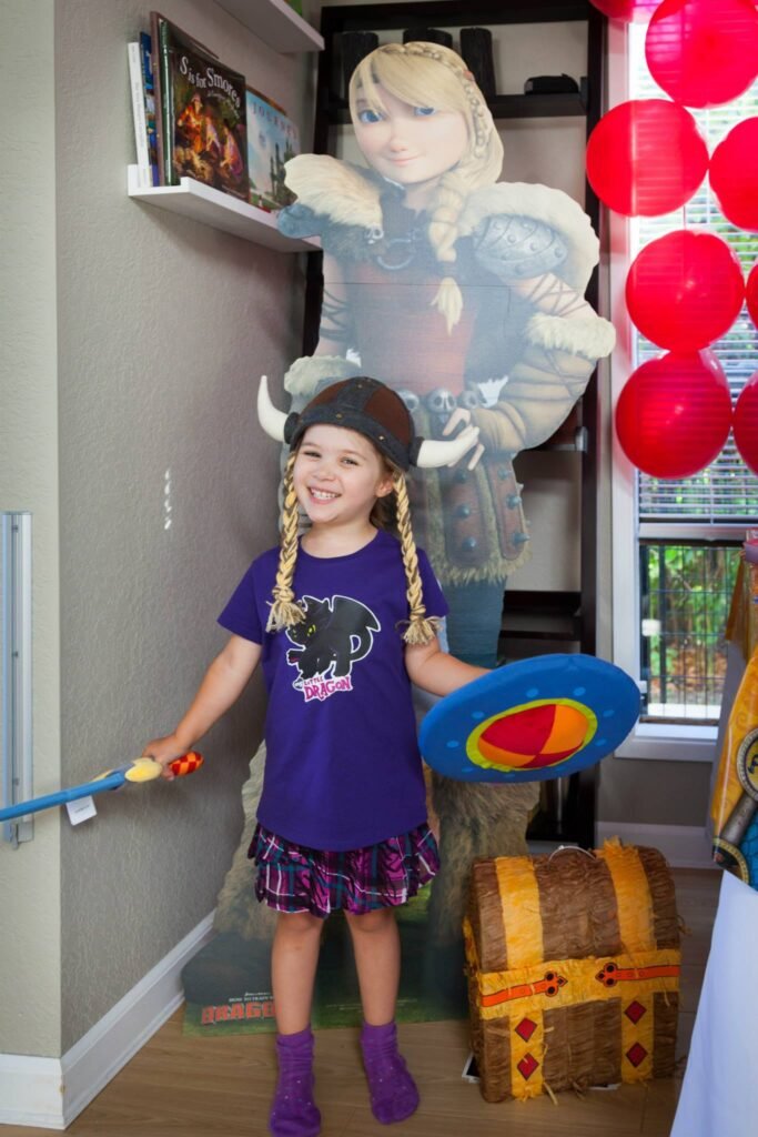 A child dressed in a Frozen costume stands beside a large character cutout and red paper lanterns. The party includes a treasure chest decoration and festive home decor in a bright room.