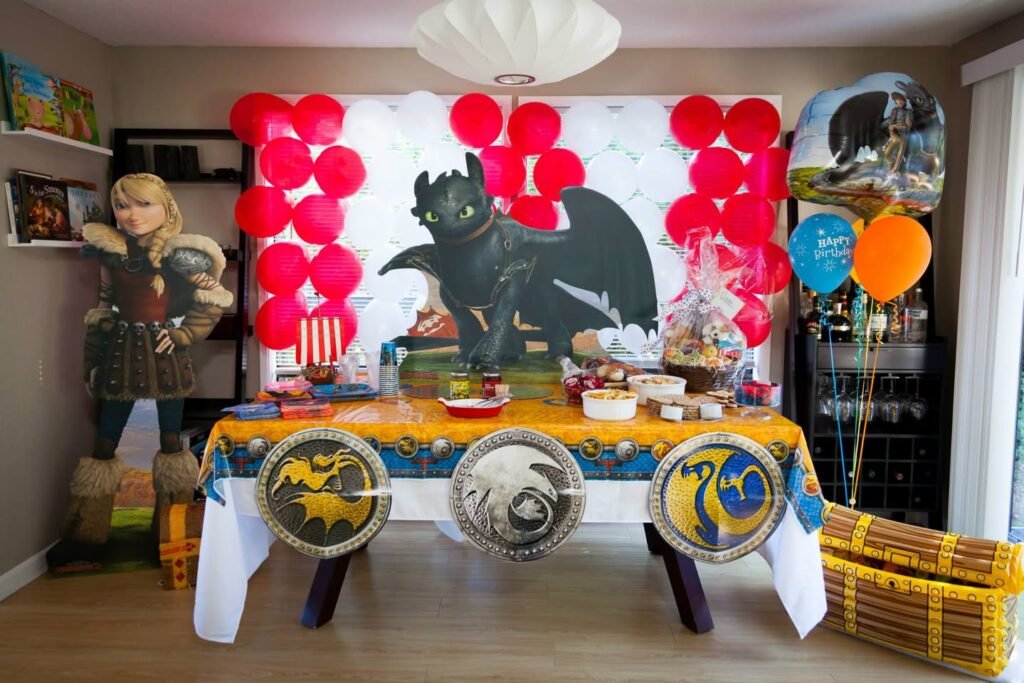 A birthday party table decorated with How to Train Your Dragon and Hiccup themed paper plates, a banner, and balloons in a living room.