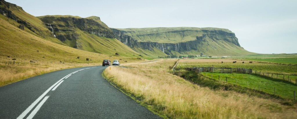 A winding asphalt road runs through grassy fields with rolling hills and ancient cliffs in the background.