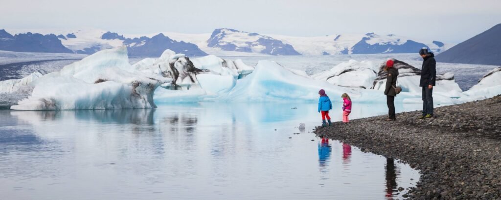 A family walks along a shoreline next to a stunning glacier lagoon with large blue icebergs floating in calm water.