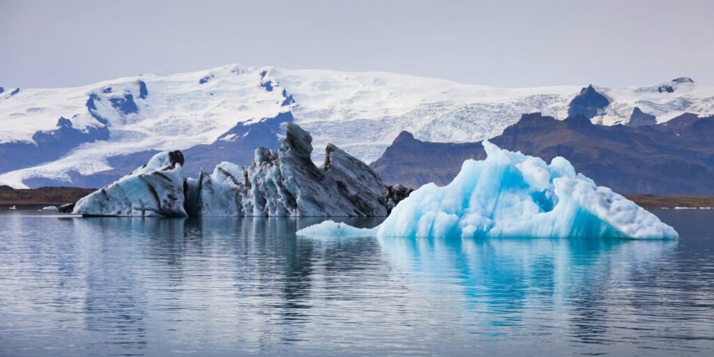Large icebergs with deep blue tones floating in a serene glacial lagoon with snow-covered mountains in the background.