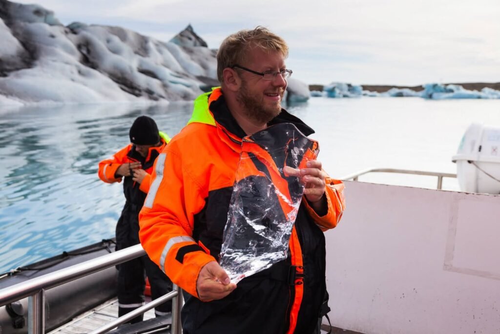 A traveler in an orange jacket holds a chunk of clear ice on a boat among glaciers.