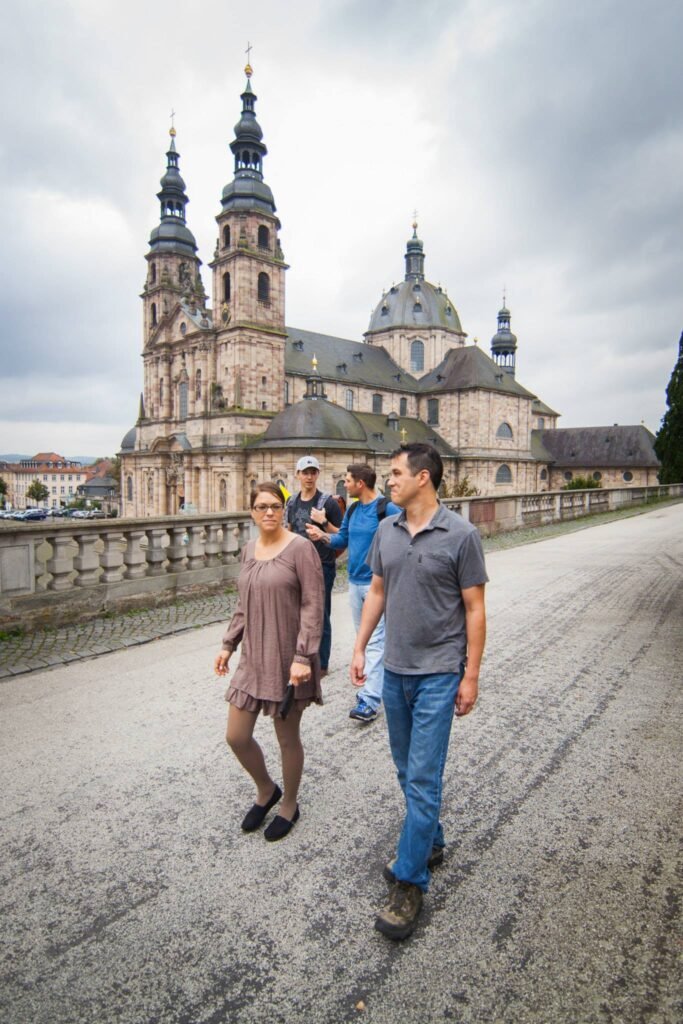 A group of travelers walks on a cobblestone street past a historical German monastery. Close-up detail of the cathedral's iconic twin spires.
