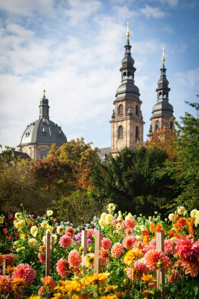 Vibrant flowers in the foreground with the iconic Berlin Cathedral towers and dome rising above the foliage against a blue sky.