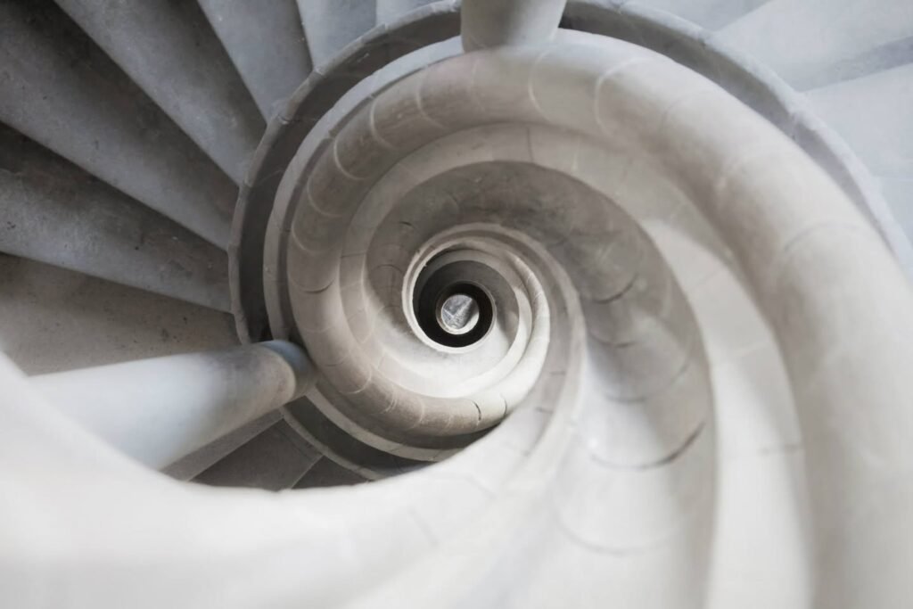 A breathtaking deep dive into a smooth, spiraling concrete staircase. The image captures the mesmerizing curvature descending into a central void.
